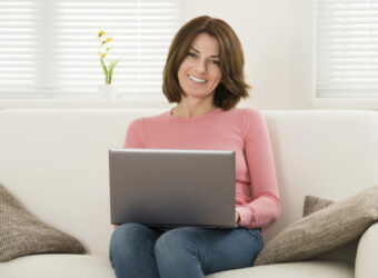 Eine lächelnde Frau sitzt mit Laptop auf einem Sofa.